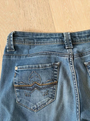 Blå bootcut jeans med broderad ficka - Säljer ett par klassiska blå bootcut jeans med snygg broderad detalj på bakfickan. Jeansen har normal midja, bälteshällor och femficksmodell. Tillverkade i denim. Det är små slitningar längs ner vid benen. 
