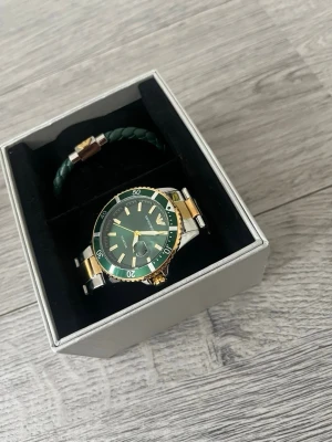 Emporio Armani grön klocka i stål - Snygg klocka från Emporio Armani med grön urtavla och grön bezel. Armbandet är i silverfärgat stål med gulddetaljer. Kommer i originalask och med matchande grön flätad armbandsaccessoar. Perfekt för dig som vill ha en stilren och exklusiv look.