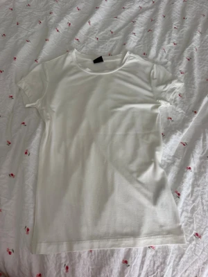 Vit basic t-shirt från Gina Tricot - En klassisk vit t-shirt från Gina Tricot i storlek XS. T-shirten har rund halsringning och korta ärmar, perfekt för en enkel och stilren look. Tillverkad i mjuk bomull som känns skön mot huden.