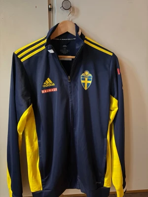 Svensk landslagsjacka Adidas blå/gul - Snygg blå och gul landslagsjacka från Adidas med Sveriges fotbollsförbunds logga på bröstet. Jackan har hel dragkedja, gula detaljer på ärmar och axlar samt Bauhaus- och Adidaslogga på framsidan. Perfekt för sena VM kvällar!