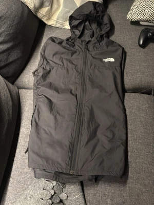 Svart vindjacka från The North Face - Svart vindjacka med huva från The North Face. Jackan har hel dragkedja framtill, två sidofickor och en vit logga på bröstet. Tillverkad i lätt och smidigt material som skyddar mot vind. Perfekt för blåsiga dagar och enkel att packa ner i väskan.