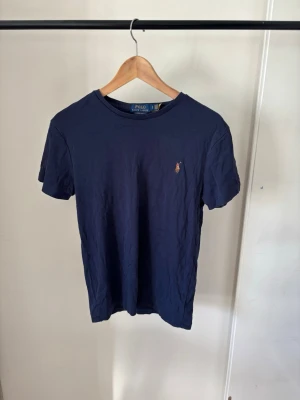 Mörkblå t-shirt från Polo Ralph Lauren - Snygg mörkblå t-shirt från Polo Ralph Lauren med klassisk rund hals och liten broderad logga på bröstet. Tillverkad i mjuk bomull och har en normal passform. Perfekt för en stilren och avslappnad look.