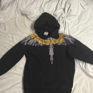 Svart hoodie Marcelo Burlon fjädermönster - Säljer en svart hoodie från Marcelo Burlon. Men har sytt den in den lite på sidorna så den sitter perfekt  i midjan för en som har S i storlek eller lite mindre. County of Milan med ikoniskt fjädermönster i gult, vitt och lila runt halsen. Hoodien har huva och är tillverkad i mjukt bomullsmaterial. Perfekt för dig som vill sticka ut med en unik design.