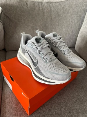 Nike Vomero 18 - Snygga grå Nike Air Zoom sneakers med svart swoosh och vita detaljer. Ovandelen är i ventilerande mesh och sulan har tydlig dämpning för extra komfort. Perfekta för dig som gillar sportig stil och vill ha riktigt sköna skor till vardagen. Helt nya och oanvända 