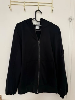 Svart hoodie med dragkedja från C.P. Company - Säljer en svart hoodie från C.P. Company med dragkedja framtill och klassisk huva. Hoodien har två fickor och den ikoniska linsen på ärmen. Perfekt för dig som gillar streetwear och vill ha något stilrent och bekvämt.