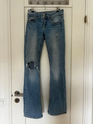 Ljusblå bootcut jeans med slitningar - Säljer ett par ljusblå bootcut jeans med slitningar och hål på ena knät. Jeansen har fem fickor, normal midja och klassisk dragkedja med knapp. Perfekta för en avslappnad och trendig look. Gissar på stlk XS