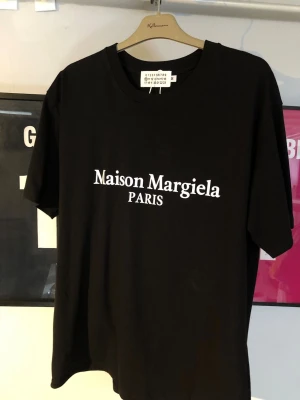 Maison Margiela svart t-shirt - Snygg svart t-shirt från Maison Margiela med klassisk vit logotyp och texten PARIS på bröstet. T-shirten har rund halsringning och korta ärmar. Perfekt för dig som gillar stilrena och ikoniska plagg.