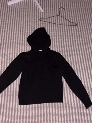 Svart stickad hoodie  - Mysig svart stickad hoodie från RYLS med huva och dragsko. Tröjan har en enkel och clean design med långa ärmar och ribbade muddar. Perfekt för dig som gillar en stilren look och vill ha något bekvämt att slänga på sig.
