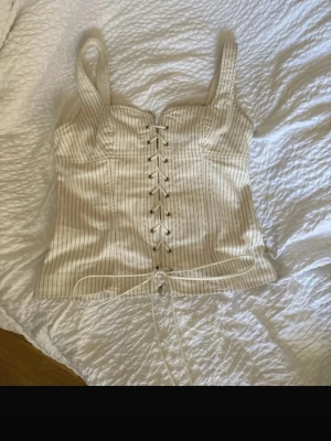 Korsettopp med snörning - Supersnygg vit korsettopp med tunna svarta ränder och snörning framtill. Toppen har breda axelband och markerad byst, vilket ger en riktigt trendig siluett. Materialet känns som bomull eller polyester och toppen har en lätt ljus ton.