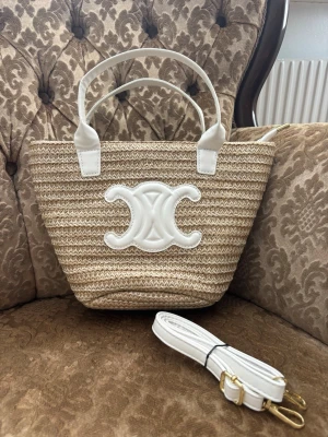 Beige stråväska med vitt emblem  - Snygg beige handväska i flätad strådesign med vita detaljer och ett stort vitt emblem framtill. 20 cm hög 30 cm bred  