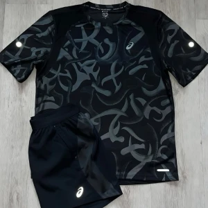 Svart träningsset från Asics - Snyggt svart träningsset från Asics med t-shirt och shorts. T-shirten har ett abstrakt mönster i grått och reflekterande detaljer på ärmar och bröst. Shortsen är enkla, svarta och har Asics-logga. Perfekt för löpning eller gym.