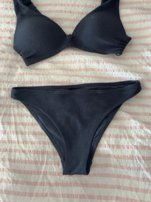 Svart bikini med ribbad struktur - Snygg svart bikini med ribbad struktur. Överdelen har vadderade kupor och breda axelband för extra stöd. Underdelen är klassisk med medelhög midja. Perfekt för strand eller poolhäng i sommar! Lappen är borta men den passar mig som är en s 