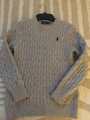 Beige kabelstickad tröja Polo Ralph Lauren - Kabelstickad beige tröja från Polo Ralph Lauren med rund halsringning och ribbade muddar. Tröjan har det klassiska broderade logotypen i svart på bröstet och är gjord i mjukt material, perfekt för kyliga dagar.