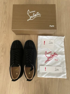 Svarta Loubs med spikar  - Svarta sneakers från Christian Louboutin med klassiska röda sulor och svarta nitar på tån. Skorna har snörning och är tillverkade i mocka med läderdetaljer. Kommer med originalkartong och dustbag. Perfekta för dig som vill sticka ut med en edgy och lyxig stil.