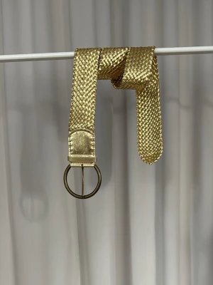 Flätat guldskärp med ring-spänne - Snyggt flätat skärp i guld med rundat ring-spänne. Skärpet har en glansig finish och passar perfekt för att lyfta vilken outfit som helst. Enkel att justera och ger en trendig touch till din stil.