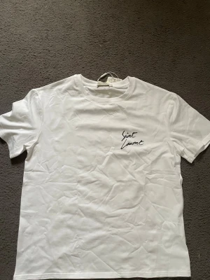 Vit Saint Laurent t-shirt - Snygg vit t-shirt från Saint Laurent med svart logotyptryck både fram och bak. Klassisk rund hals och korta ärmar. Tillverkad i mjuk bomull för en skön känsla och clean look.