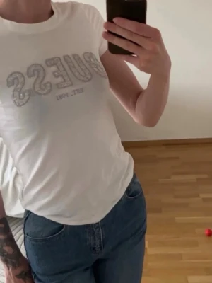 Vit kortärmad t-shirt från Guess - Säljer en vit t-shirt från Guess med korta ärmar och rund hals. Framtill står det 'GUESS EST. 1981' i glittriga bokstäver. Perfekt till jeans eller kjol och har en klassisk passform. Materialet känns mjukt och skönt, troligen bomull.