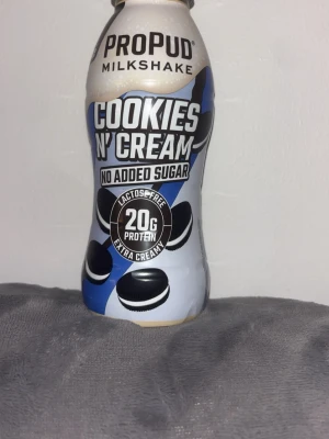 ProPud Milkshake Cookies n' Cream - ProPud Milkshake med smaken Cookies n' Cream. Innehåller 20g protein, är laktosfri och har ingen tillsatt socker. Flaskan är vit och blå med svarta kakmotiv och text i svart och vitt. Perfekt för dig som vill ha ett krämigt och gott mellanmål.