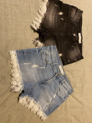 Två jeansshorts med spetskant - Säljer två par snygga jeansshorts, ett par i ljusblå denim och ett par i svart tvättad denim. Båda har slitna detaljer och vit spetskant längst ner. Perfekta för sommaren och ger en cool, feminin vibe till din outfit.