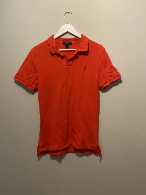 Röd pikétröja från Polo Ralph Lauren - Säljer en klassisk röd pikétröja från Polo Ralph Lauren med blå broderad logga på bröstet. Tröjan har korta ärmar, krage och knappar vid halsen. Perfekt till sommaren och enkel att matcha med jeans eller shorts. Tröjan sitter som en strl Xs/s