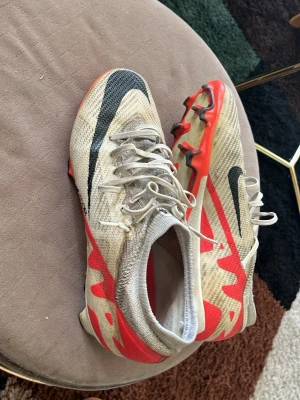 Nike fotbollsskor beige/röd - Säljer ett par Nike fotbollsskor i beige med röda och svarta detaljer. Skorna har snörning, låg profil och en ovandel i mesh-liknande material för bra ventilation. Sulan har dobbar för spel på gräs. Perfekta för dig som vill ha stilrena och lätta skor på planen.
