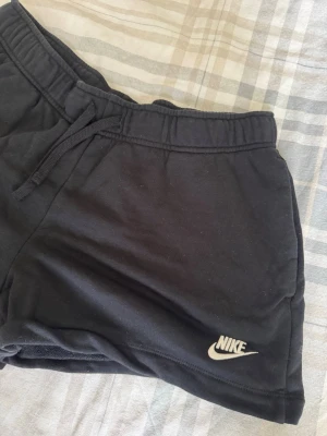 Svarta shorts från Nike med snörning - Säljer ett par svarta shorts från Nike med klassisk logga broderad i vitt på benet. Shortsen har elastisk midja med snörning för skön passform och är gjorda i mjuk bomull. Perfekta för chill dagar eller träning.