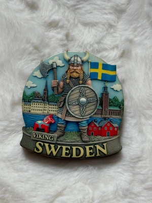 Viking Sweden kylskåpsmagnet - Dekorativ kylskåpsmagnet med vikingamotiv, svensk flagga och klassiska svenska byggnader. Färgglad design med inslag av rött, blått, gult och grått. Perfekt souvenir eller present till någon som gillar Sverige och vikingar.