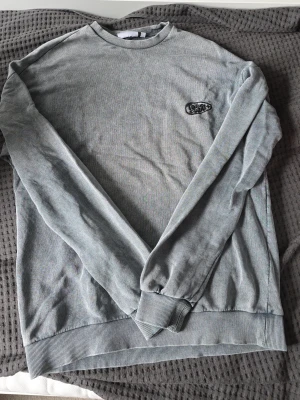 Ljusgrå sweatshirt med broderad logga ffån junkyard - En stilren ljusgrå sweatshirt med rund hals och ribbade muddar. Tröjan har en diskret broderad logga på bröstet och är tillverkad i mjukt material som passar perfekt till vardagslooken. Långärmad modell med relaxed fit. Passar s och m. Junkyard