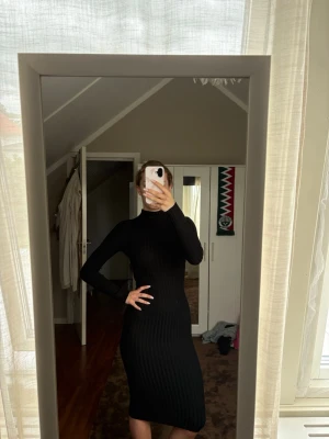Svart ribbad långärmad klänning - Säljer en svart, ribbad klänning med lång ärm och hög hals. Klänningen är figurnära och har en klassisk, stilren look som passar perfekt till många tillfällen. Materialet är stretchigt och bekvämt, vilket ger en snygg passform.