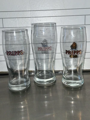 Pripps ölglas 80kr st - Tre klassiska ölglas från Pripps med tryckt logga och skeppsmotiv. Glasen är genomskinliga och har en lätt böjd form som passar perfekt till öl eller andra drycker. Snygga att ha i köksskåpet eller till samlingen.