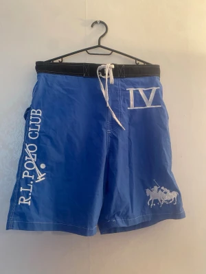 Blå badbyxor från R.L. Polo Club - Snygga blå badbyxor från R.L. Polo Club med vita broderade detaljer, inklusive klubbens logga och polospelare. De har svart midjeresår med vit dragsko och tryck i vitt på benet. Perfekta för stranden eller poolen.