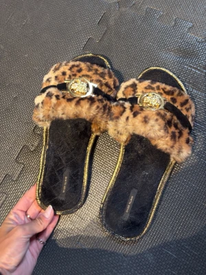 Leopardmönstrade fluffiga tofflor - Säljer ett par snygga tofflor från River Island med leopardmönstrad fuskpäls, guldfärgade detaljer och dekorativa spännen framtill. Sulan har en glittrig guldkant och innersulan är svart. Perfekta för dig som vill ha något extra till hemmastilen.