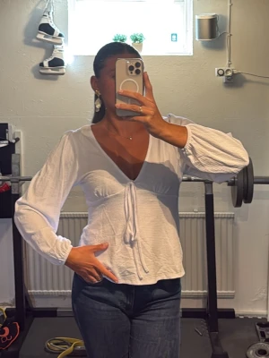 Vit trending topp med v-ringning - Säljer en vit omlottopp med v-ringning. Toppen har långa, lätt puffade ärmar och är gjord i ett tunt, mjukt material som känns luftigt. Perfekt att styla med jeans eller kjol för en old money lock!