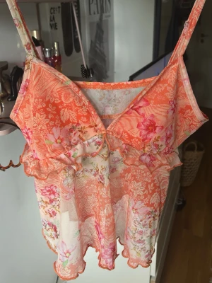 Blommig mesh body med volanger - Söt body i tunn mesh med blommigt mönster i orange, rosa och vitt. Spaghettiband och volangdetaljer längs bysten och nederkanten ger en romantisk vibe. Perfekt för dig som gillar feminina detaljer och lätta material.