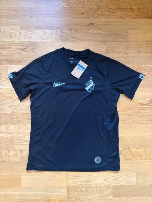 Svart AIK matchtröja Nike - Snygg svart matchtröja från Nike med AIK-logga på bröstet och diskreta detaljer i ljusblått. Tillverkad i ett lätt och snabbtorkande funktionsmaterial, perfekt för träning eller match. Klassisk passform med korta ärmar och rund hals.