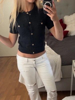 Svart kortärmad topp med knappar - Snygg svart topp med korta ärmar och dekorativa guldknappar framtill. Toppen är croppad och har två fejkade bröstfickor. Perfekt till jeans eller kjol för en trendig look.