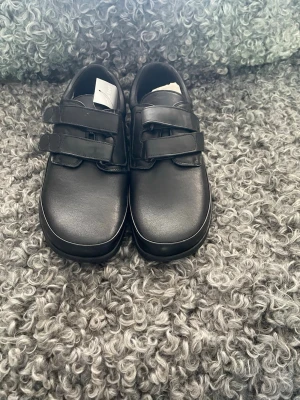 Svarta loafers med kardborreband - Säljer ett par svarta loafers med två breda kardborreband över foten. Skorna har en stilren och enkel design med rundad tå och låg sula. Materialet ser ut att vara slätt konstläder som ger en clean look. Perfekta för dig som gillar klassiska och bekväma skor.
