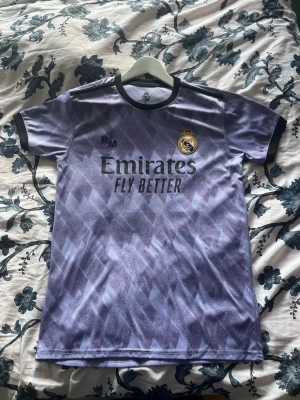 Lila Real Madrid matchtröja Benzema - Säljer en lila Real Madrid matchtröja med svart krage och ärmslut. Tröjan har klubbmärket och sponsortryck fram samt Benzema och nummer 9 på ryggen. Tillverkad i ett lätt och ventilerande material, perfekt för fotboll eller träning.