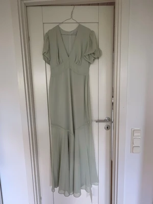 Ljusgrön långklänning med puffärm - Elegant långklänning i ljus saige (ljusgrön) med v-ringning och puffärmar. Klänningen har en mjuk, flytande siluett och är tillverkad i ett tunt, lätt material som ger ett romantiskt intryck. Perfekt för sommarens alla tillfällen.