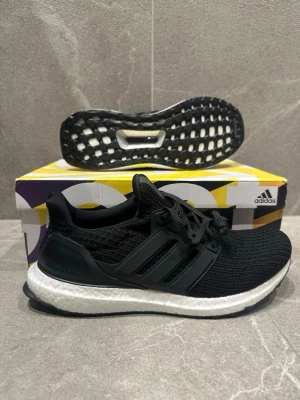 Adidas Ultraboost svarta sneakers - Svarta Adidas Ultraboost sneakers med stickad ovandel i mesh och klassiska tre ränder på sidan. Skon har en vit Boost-mellansula för extra komfort och svart yttersula med grepp. Perfekt för dig som gillar stilrena och sportiga sneakers.