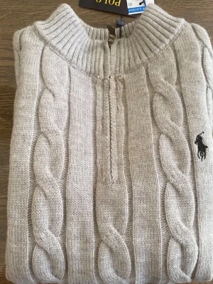 Ljusgrå kabelstickad tröja Polo Ralph Lauren - Säljer en ljusgrå kabelstickad tröja från Polo Ralph Lauren med halv dragkedja och hög krage. Tröjan har det klassiska broderade logotypen på bröstet och ribbade muddar vid ärmslut och nederkant. Perfekt för dig som gillar stilren och tidlös design.