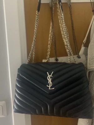 Svart YSL handväska i läder - Säljer en svart handväska från Saint Laurent med klassisk YSL-logga i silver framtill. Väskan är quiltad i chevronmönster och har en kedjerem i silver med läderdetaljer. Perfekt för dig som vill ha en lyxig och tidlös accessoar.
