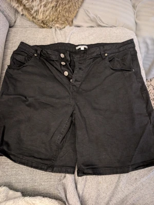 Svarta  Stretch shorts med knappar stl 46 - Svarta shorts i loose fit med hög midja och knappgylf. Klassisk femficksmodell med bälteshällor och stora bakfickor. Perfekta för en avslappnad stil och lätta att matcha med olika toppar. 50 cm rakt över midjan
