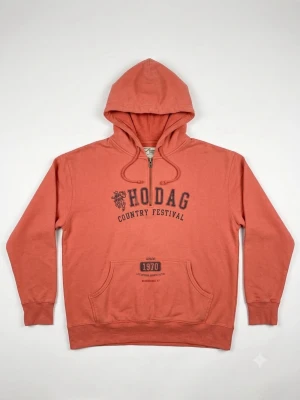 Korallfärgad Hodag hoodie Artisans - Snygg korallfärgad hoodie från Artisans med trycket 'Hodag Country Festival' på bröstet. Klassisk känguruficka framtill, dragsko i huvan och ribbade muddar. Tillverkad i 80% bomull för en mjuk och skön känsla. Perfekt för dig som gillar festivalvibbar.