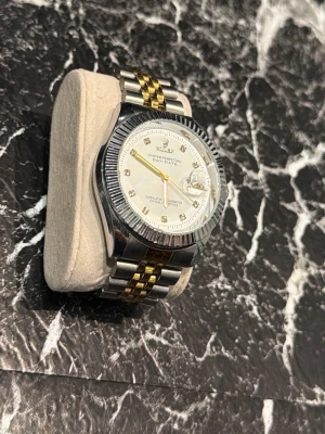 Silver och guld armbandsur - Stilren klocka med tvåfärgat armband i silver och guld. Urtavlan är vit med markerade index och datumvisning. Boetten är rund och har en räfflad kant. Passar perfekt för dig som vill ha en klassisk accessoar med lyxig känsla.