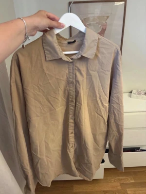 Beige långärmad skjorta - Säljer en enkel och stilren beige skjorta med klassisk krage och knäppning framtill. Skjortan har långa ärmar och är tillverkad i ett mjukt bomullstyg. Perfekt att styla lager på lager eller bära som den är.