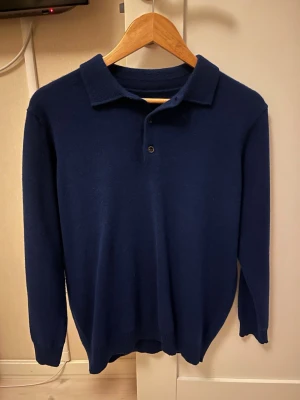 Mörkblå cashmerepiké  - Snygg mörkblå pikétröja i mjuk cashmere med lång ärm och klassisk krage. Tröjan har tre knappar framtill och ribbade muddar vid ärmslut och nederkant. Perfekt för dig som gillar stilrena och bekväma plagg. Blå Pike tröja i cashmere, stilren passar till allt! Finns inte någon etiker i tröjan så inte säker på vilket märke det är