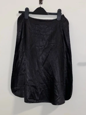 Svart kortkjol i satin från H&M - En svart kortkjol i glansigt satinmaterial från H&M. Kjolen har en enkel och stilren design med lätt A-linjeformad siluett. Perfekt att matcha med en crop top eller stickad tröja för en trendig look.
