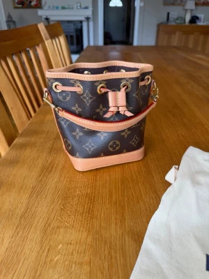 Louis Vuitton monogram bucket bag - Säljer en liten bucket bag från Louis Vuitton i klassiskt monogrammönster. Väskan är brun med beige och guldiga detaljer, har dragsko upptill och en kort axelrem. Tillverkad i canvas med läderdetaljer. Så fin och nästan som ny! Modellen ”nano noé” har NFC tagg se sista bilden.