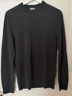 Mörkgrå stickad tröja från H&M premium - En stilren mörkgrå stickad tröja från H&M premium med rund halsringning och långa ärmar. Tröjan har ribbade muddar vid hals, ärmslut och nederkant. Perfekt för lager på lager och enkel att matcha med olika outfits.